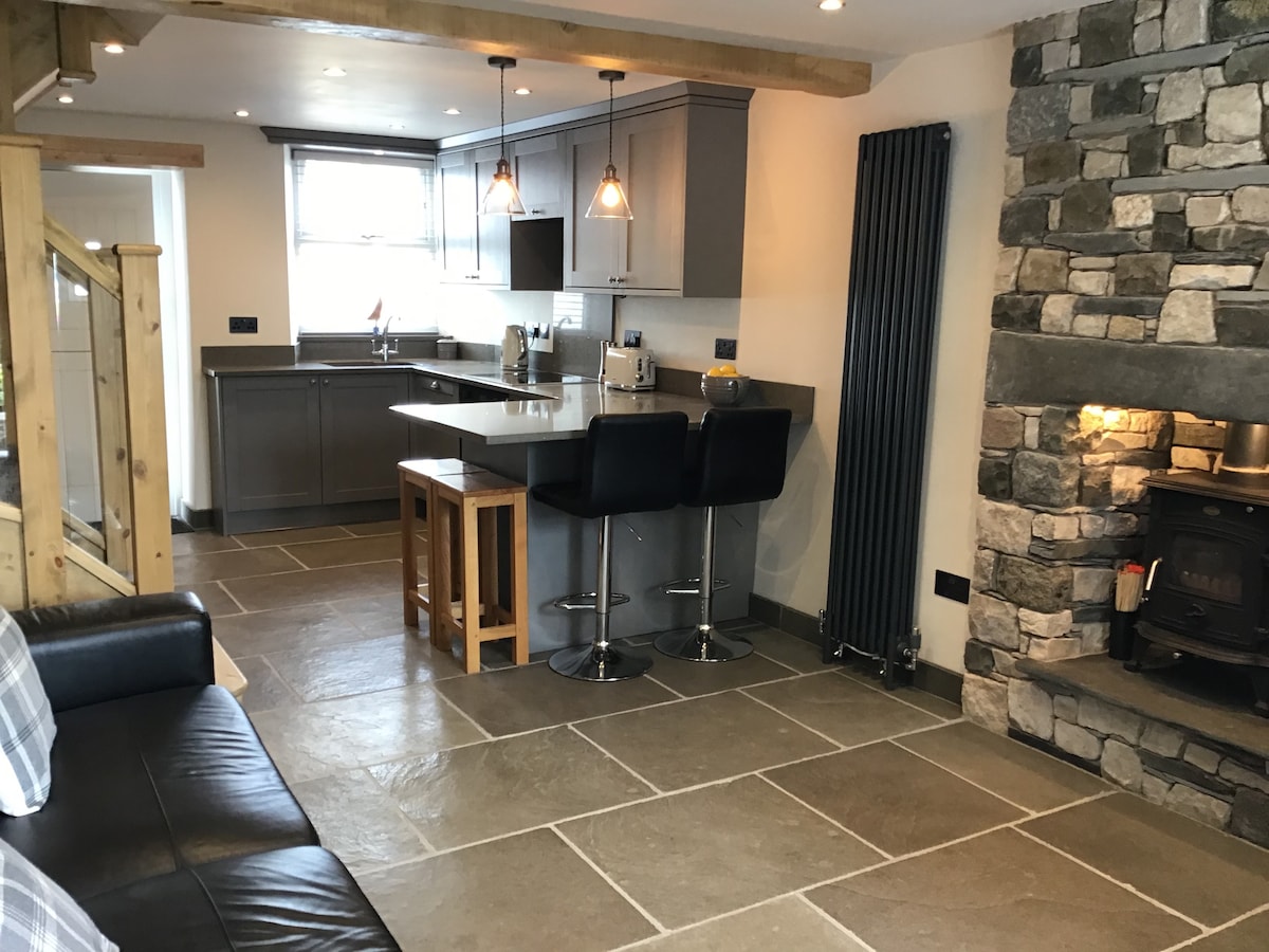 An open-plan kitchen and dining area is highlighted by grey cabinetry and quartz countertops. Bar stools are positioned at a central island, while a stone fireplace with a log burner adds warmth to the living space. Underfloor heating enhances comfort throughout.
