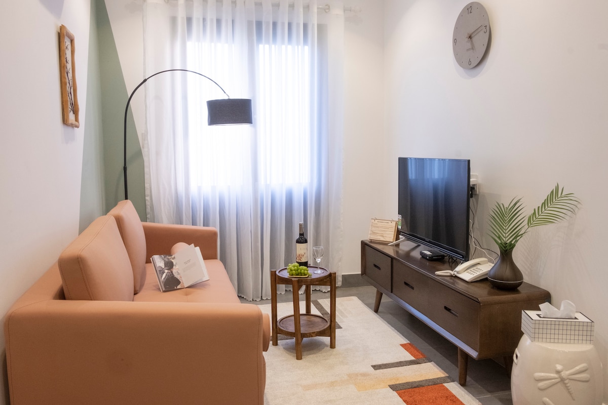 A cozy living area features a modern sofa, a wooden coffee table with snacks, and a flat-screen TV. Natural light filters through sheer curtains on a window, and a wall clock adds a touch of functionality. Decorative elements include a potted plant and a floor lamp.