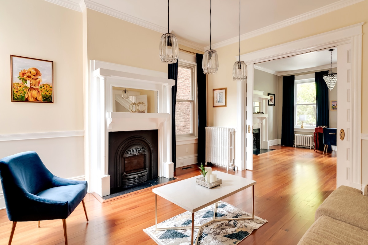 A spacious living area features hardwood flooring and high ceilings, complemented by large windows allowing natural light. A decorative fireplace is framed by a stylish mantel, while a coffee table rests on an area rug. Modern light fixtures hang from the ceiling, adding elegance to the space.