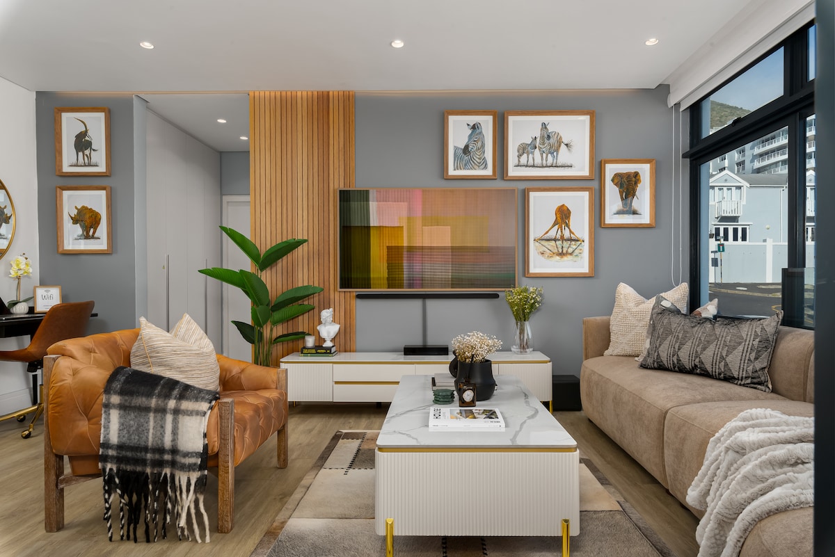The living area showcases an open layout with a mix of contemporary furniture and artwork. A comfortable sofa and an accent chair are positioned around a central coffee table. Nature-inspired decor, including a potted plant, complements the light wood and neutral color palette.