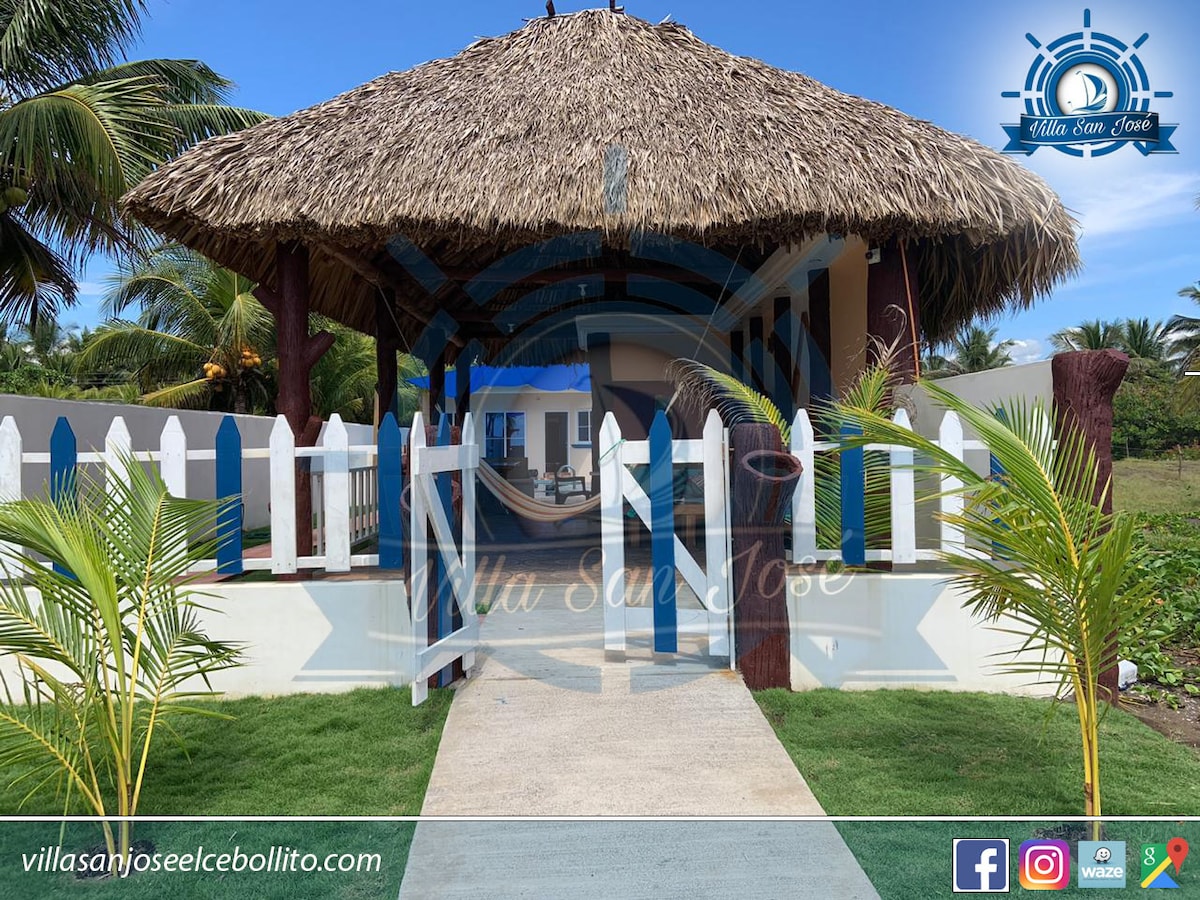 An inviting entrance features a traditional thatched-roof structure, framed by vibrant greenery and a white picket fence. A smooth pathway leads to a welcoming outdoor space, designed for relaxation and socializing.