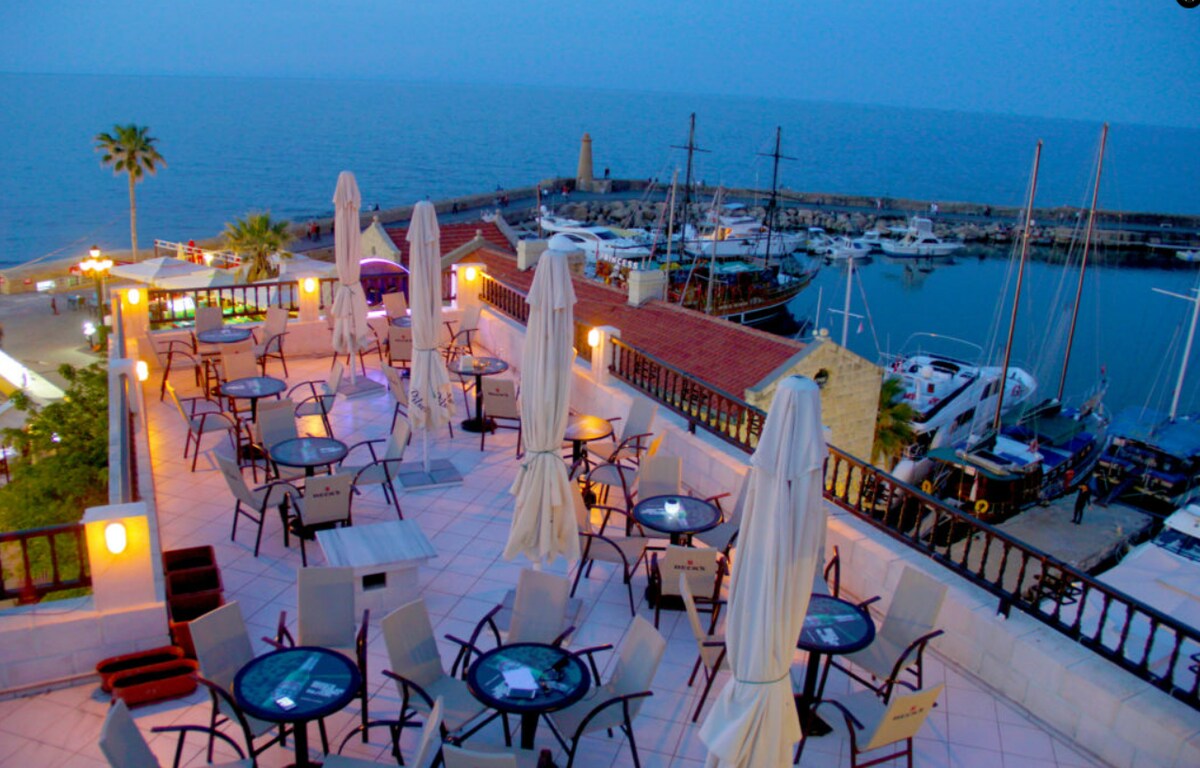 An elevated terrace is visible, featuring round tables and chairs arranged beneath white umbrellas. The sea and a distant harbor filled with boats stretch out in the background. Soft lighting highlights the space, creating a calm ambiance as dusk settles.
