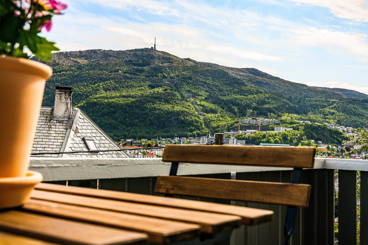 A wooden table and chair are positioned on a balcony, with a backdrop of a mountain covered in lush greenery. A flowerpot with vibrant blooms sits in the foreground, adding a touch of color to the scenic view of the distant mountain peak.