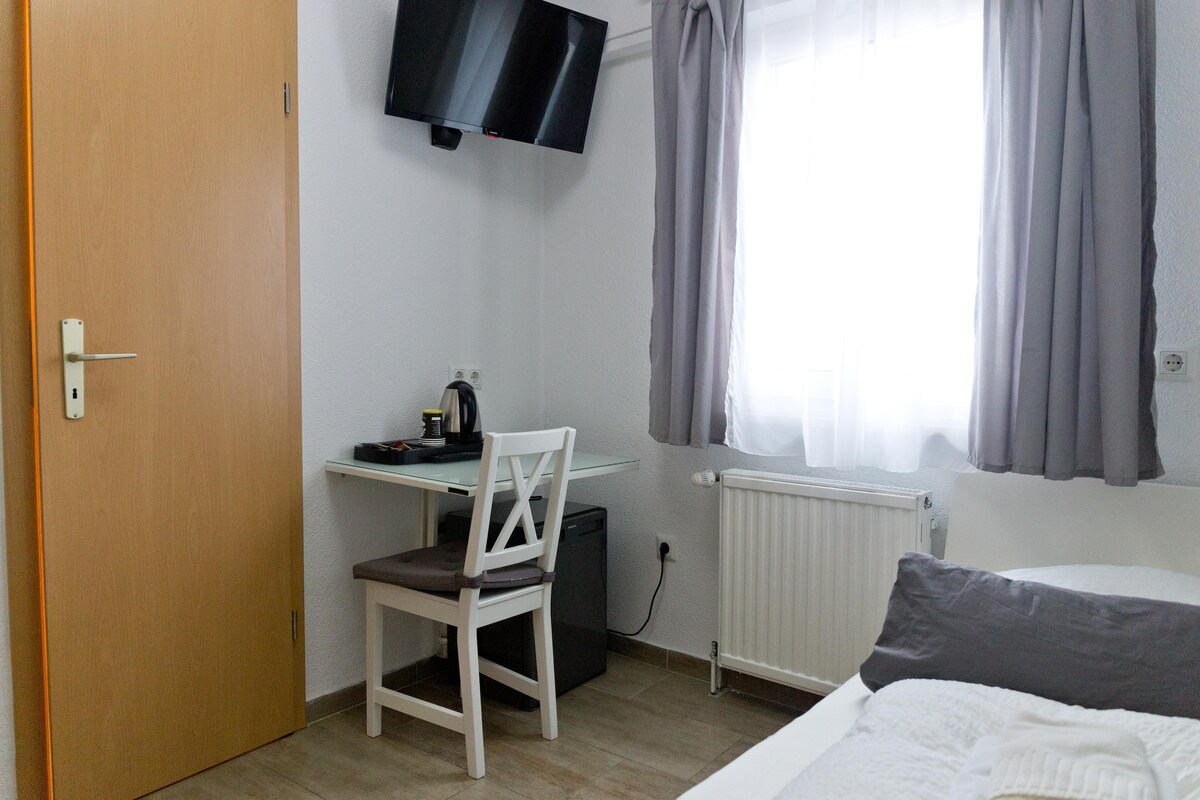 A compact room is equipped with a flat-screen TV mounted on the wall. A desk with a chair is positioned next to a window, which is dressed with light curtains. A kettle and a small fridge are visible on the side, offering convenience for guests.