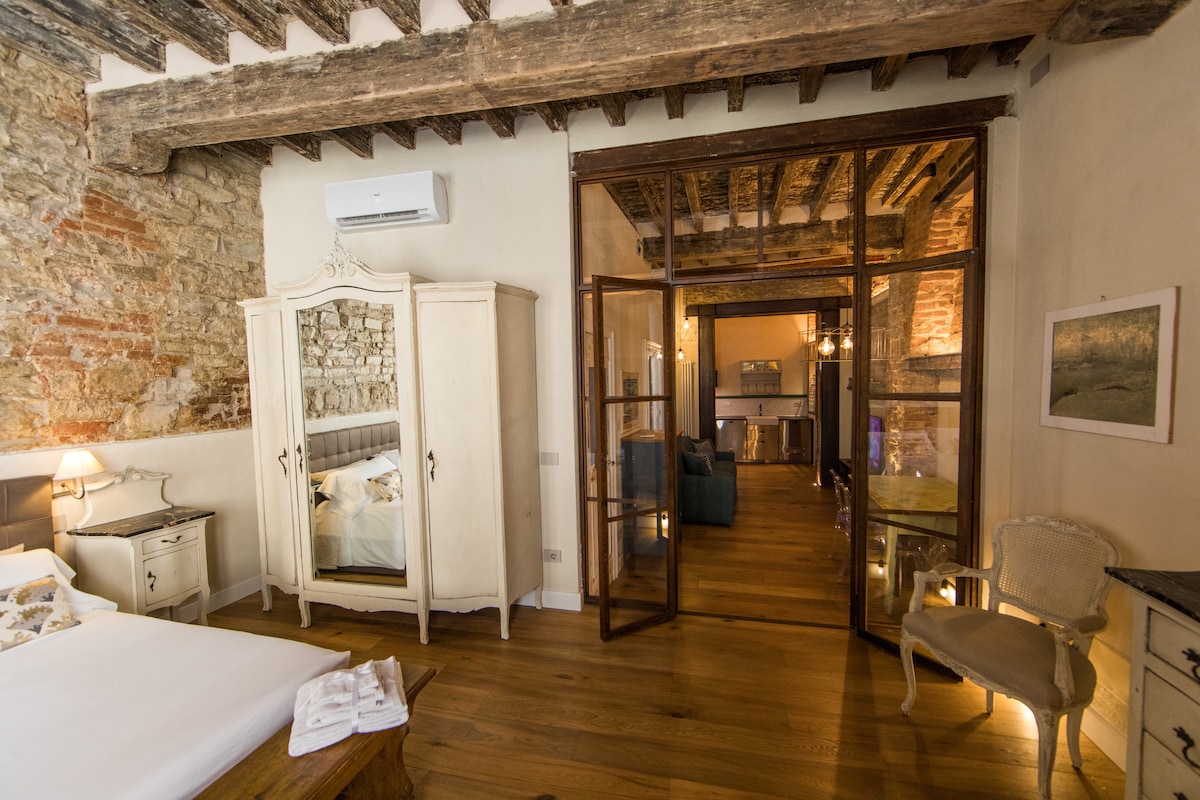 A spacious bedroom features a double bed with crisp white linens. An antique wardrobe stands against a textured stone wall, while wooden beams add character to the ceiling. The room opens to a living area through glass doors, with natural light enhancing the warm wood flooring.