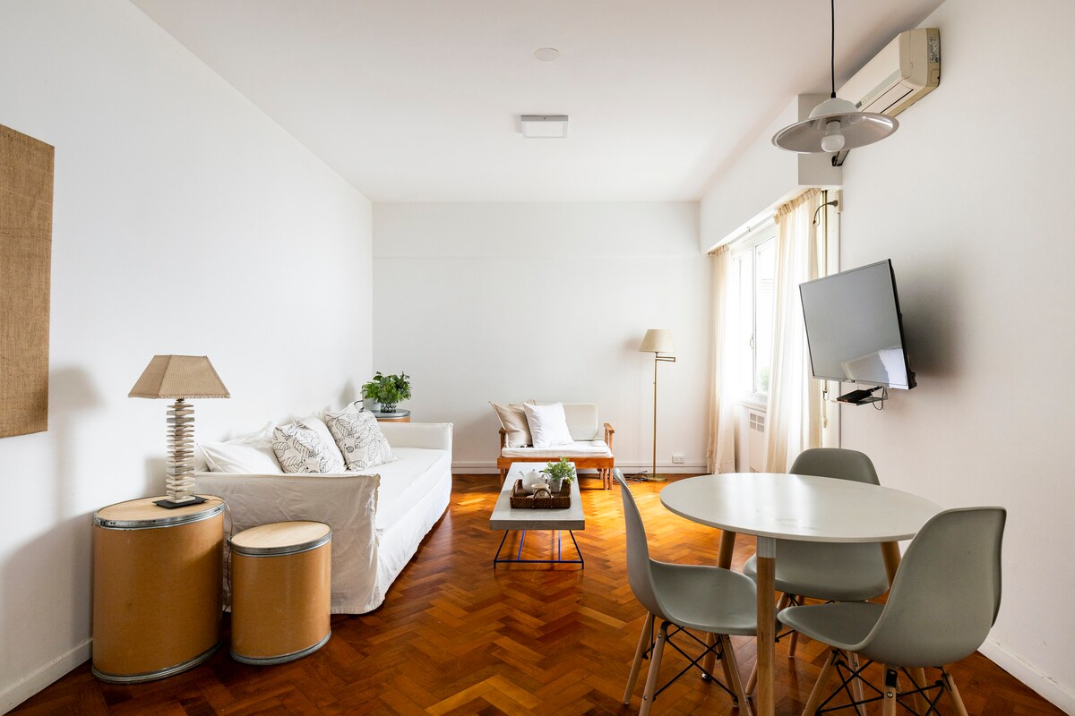 A bright living and dining area features a spacious white sofa, accompanied by a small side table and decorative plant. A round dining table with four chairs is positioned near a window that allows natural light. A mounted TV is visible on the wall, and air conditioning is present.