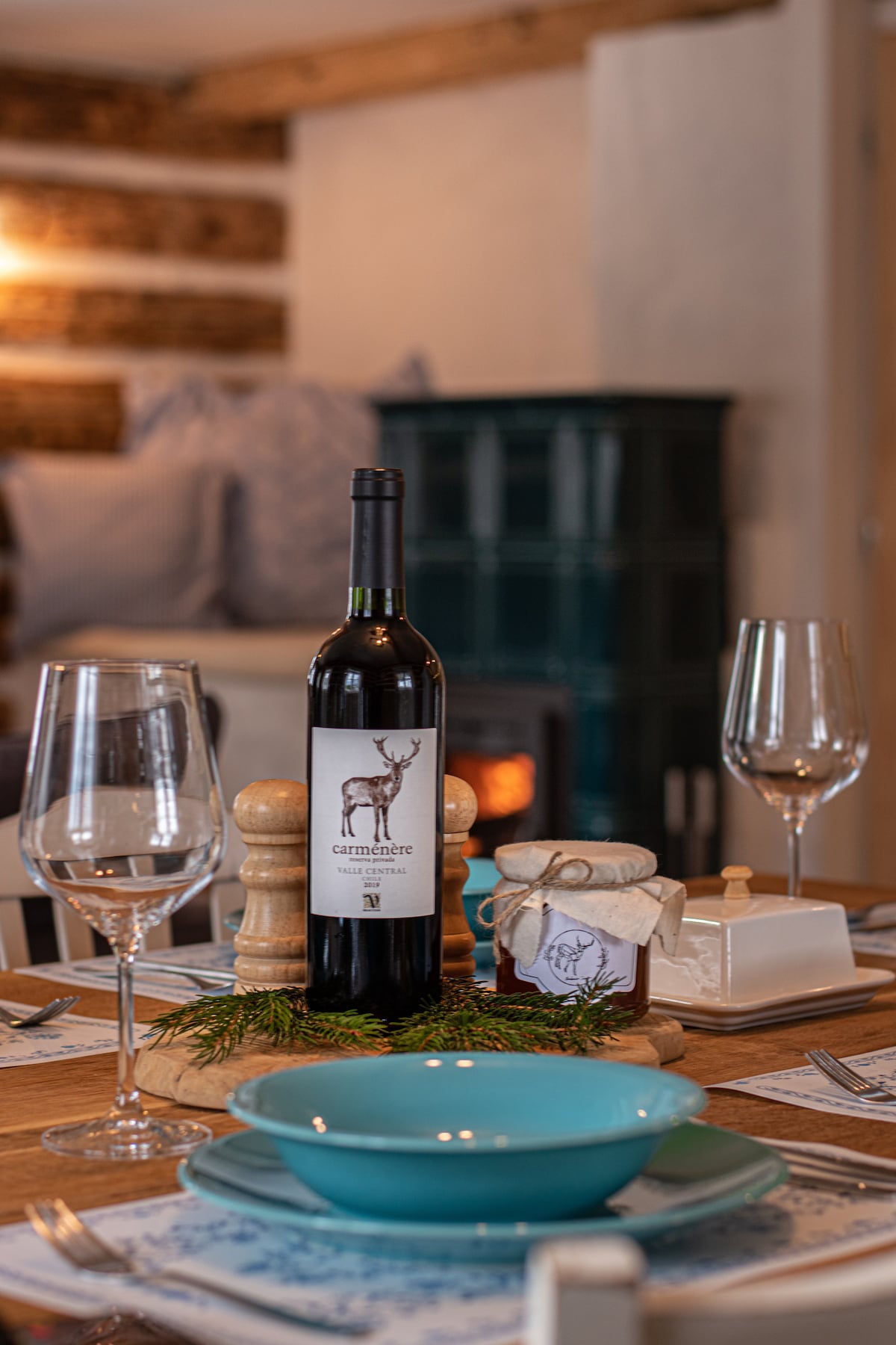 A dining table is set with elegant glassware, polished utensils, and a blue bowl at the center. A decorative wine bottle with a label featuring a deer stands alongside a small jar and butter dish. In the background, a cozy wood stove is visible, creating a warm ambiance.