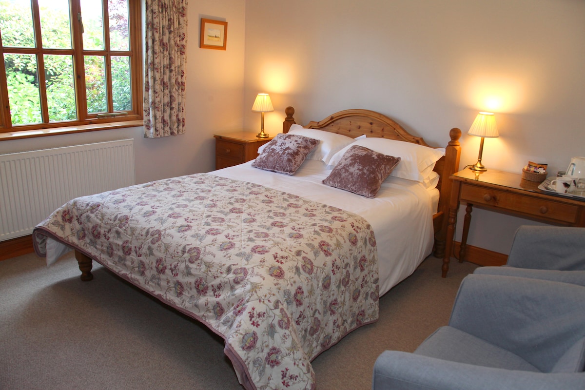 A cozy bedroom features a king size bed with a floral-patterned spread and two pillows. Soft lighting from bedside lamps illuminates the room. A chair is positioned nearby, and a window offers a view of the garden outside.