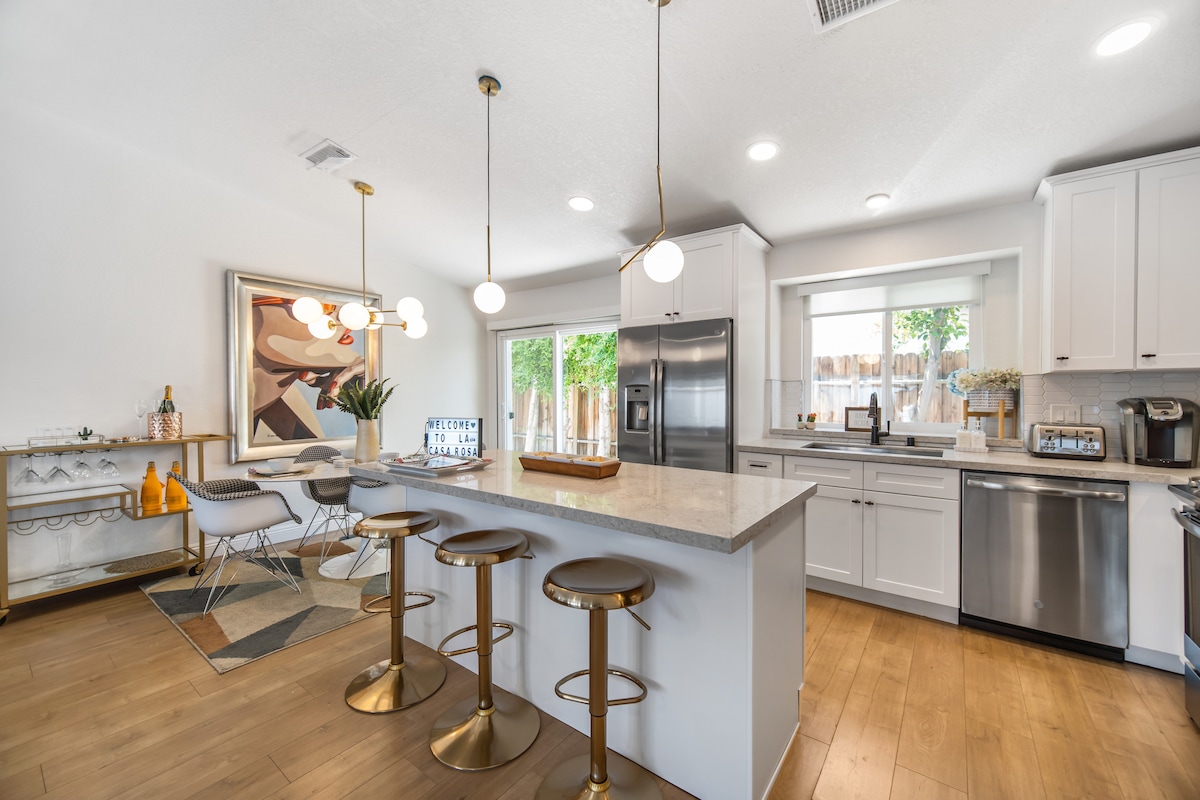 A contemporary kitchen features an expansive island with three gold-accented stools. Modern stainless-steel appliances are positioned along the walls, alongside a farmhouse sink. Natural light fills the space through a window, while a colorful artwork adds a touch of elegance to the room.