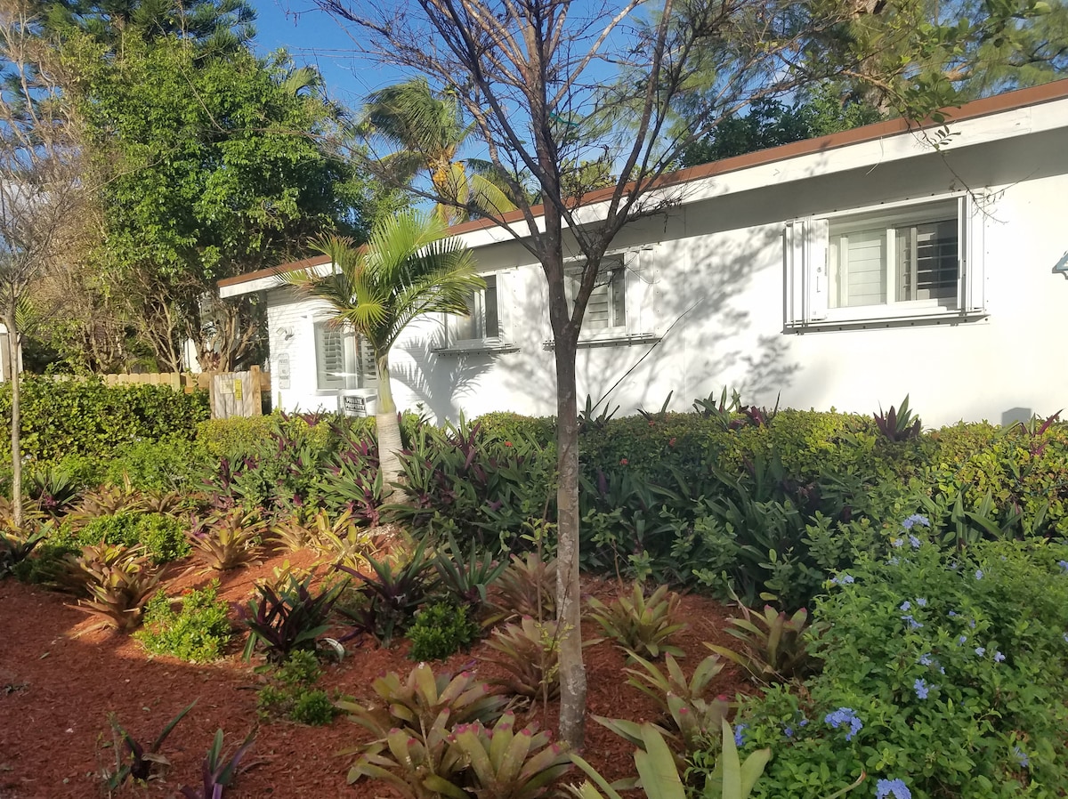 The exterior of the unit is surrounded by lush, vibrant landscaping featuring a variety of tropical plants. A well-maintained garden bed with reddish-brown mulch provides color contrast, while the building's windows are framed by greenery, creating a serene and inviting entrance.