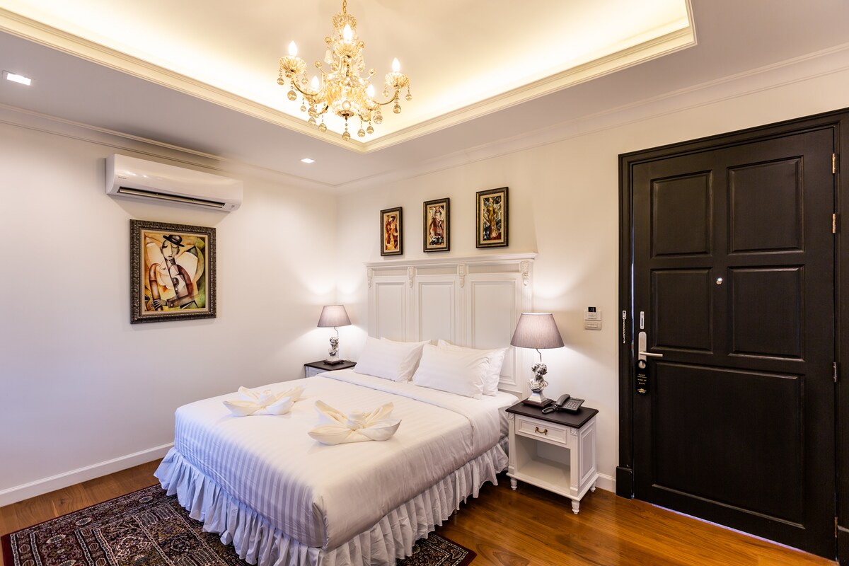 The room features a spacious bed adorned with decorative towels, complemented by elegant bedside lamps and artwork on the walls. A chandelier hangs from the ceiling, while a wooden door adds a touch of sophistication to the welcoming space.