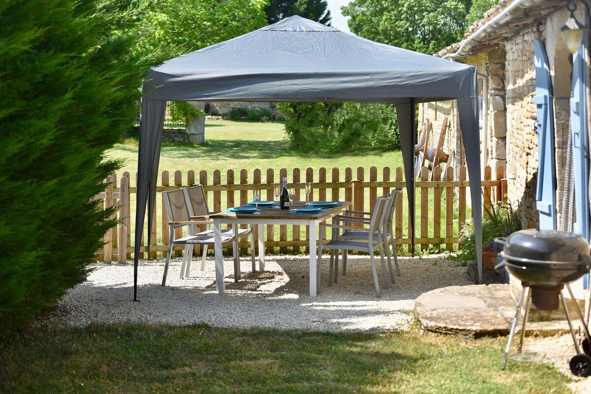A shaded outdoor seating area is displayed under a canopy, featuring a table set with chairs. A BBQ grill is positioned nearby. Lush greenery and a wooden fence create a private atmosphere in the surrounding garden.