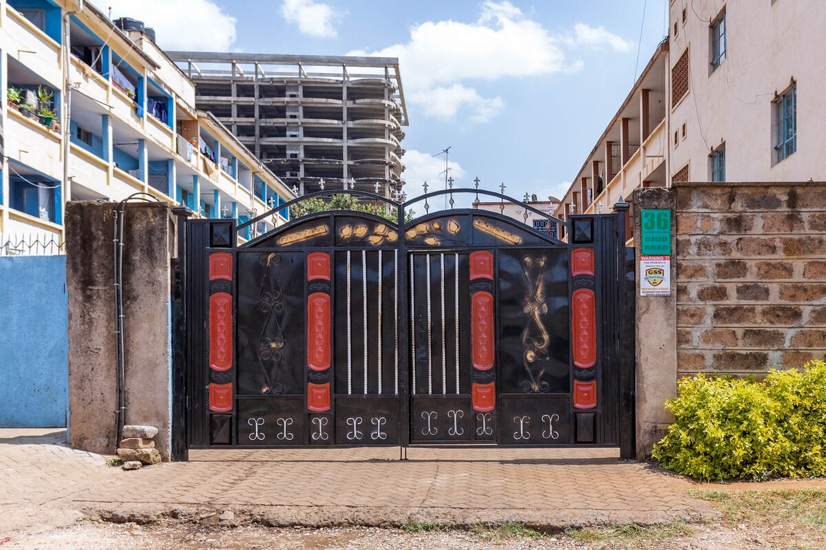 An ornate black gate is showcased, featuring red accents and intricate designs. Surrounding structures include low-rise buildings, with trees and shrubbery visible nearby. The sky above reveals fluffy clouds, hinting at a pleasant day. The entrance is flanked by a dirt path and paved areas.