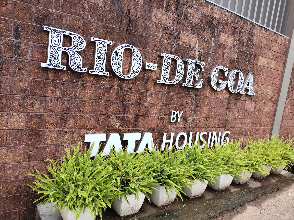 The entrance sign for Rio De Goa is prominently displayed on a textured brown brick wall. The sign features decorative lettering and is accompanied by green potted plants, adding a touch of nature to the welcoming entrance.