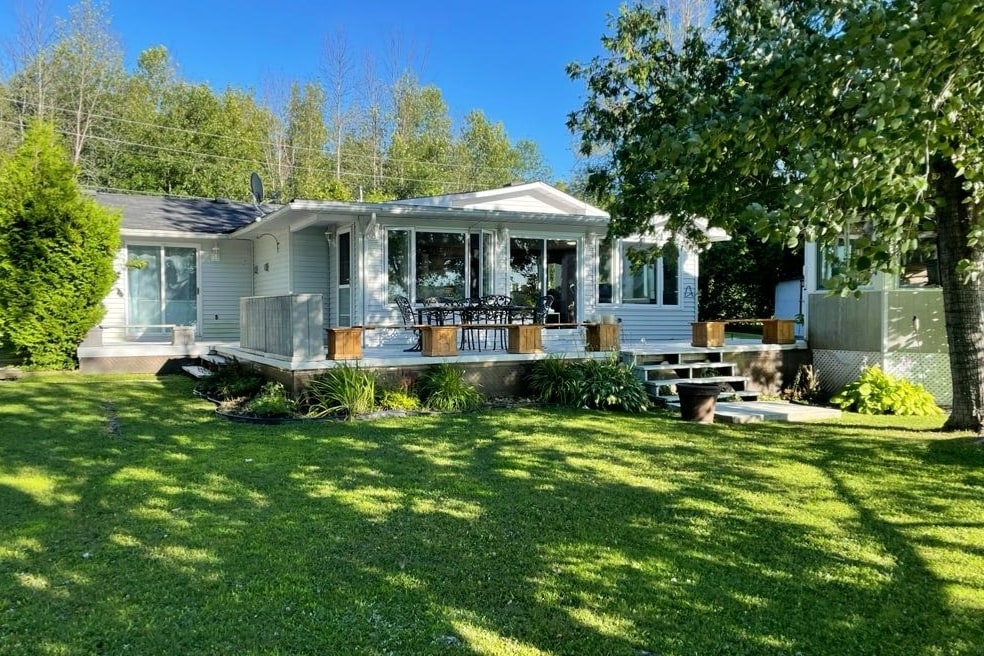 The cottage exterior showcases a well-maintained lawn with lush greenery and several trees. The large deck is featured with outdoor seating, offering a space to enjoy the surrounding nature. Sunlight filters through the trees, creating a serene and inviting atmosphere.