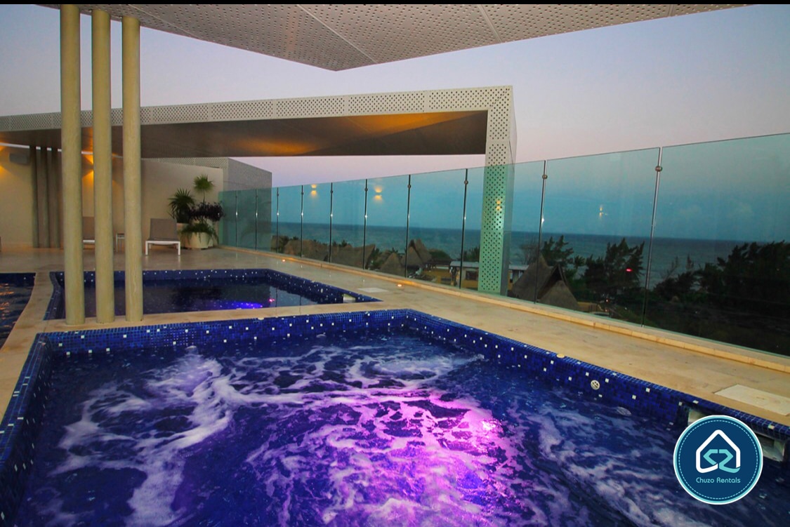 A serene rooftop area features a heated jacuzzi surrounded by glass railings, providing an unobstructed view of the ocean. The water displays a soft purple hue, enhancing the tranquil ambiance. Sun loungers are positioned nearby, inviting relaxation in the warm atmosphere.
