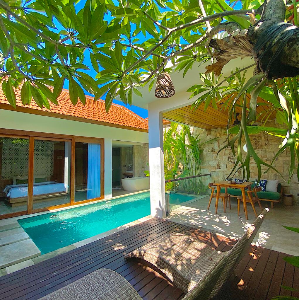An outdoor seating area is displayed alongside a private pool, accented by lush greenery. Large glass doors lead to a guest room featuring visible interior elements. Natural light brightens the space, highlighting the seamless connection between indoor and outdoor areas.