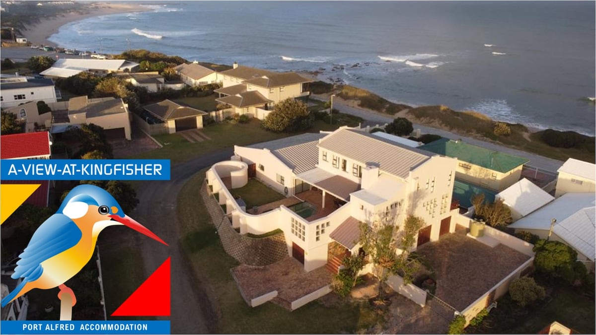 Aerial view of the accommodation reveals a spacious property surrounded by greenery, with proximity to a coastline featuring gentle waves. The well-maintained building displays multiple sections, with a mixture of roofs creating an appealing architectural design.