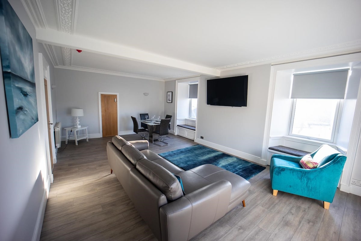 A spacious lounge area is shown, featuring a large, comfortable sofa in a neutral tone and an eye-catching teal chair. Natural light flows through the windows, illuminating the modern décor, which includes a flat-screen television and a dining table in the background.