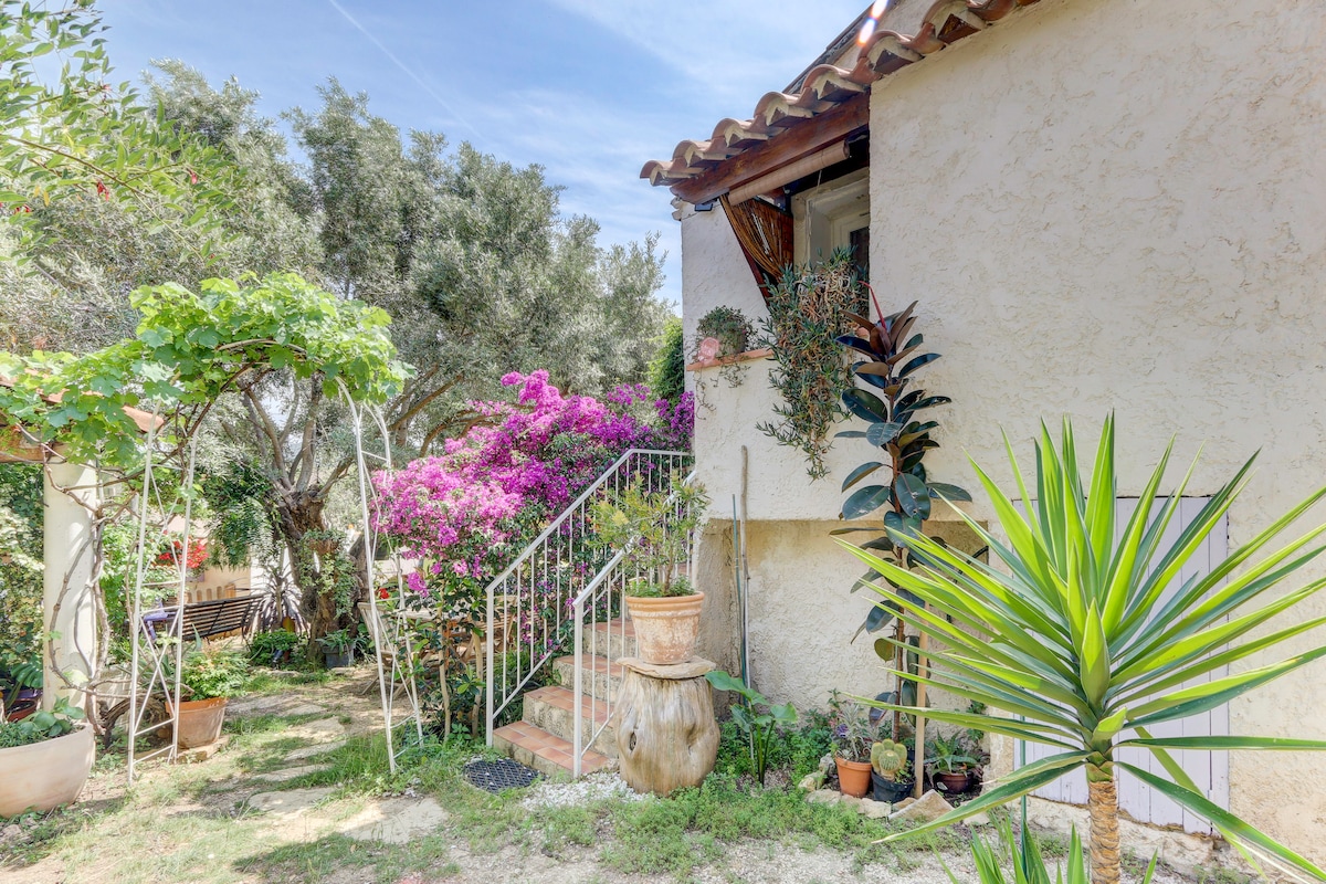 A charming exterior view of the studio features a well-kept garden with lush greenery and vibrant pink bougainvillea. A stone staircase leads to the entrance, flanked by decorative potted plants and a small stone fountain, contributing to a welcoming ambiance.