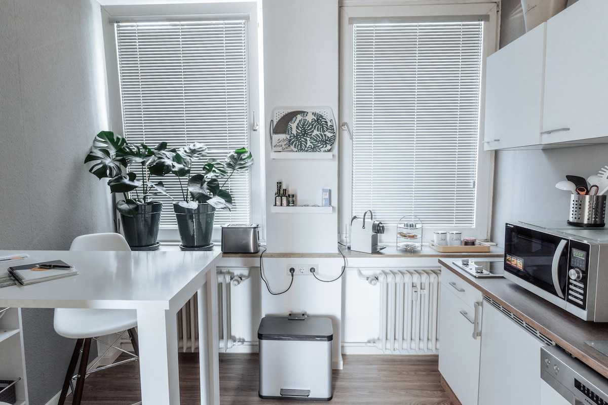 A modern kitchen is depicted with two large windows allowing natural light. A high table with bar stools is seen alongside a microwave and a silver kitchen appliance. Potted plants add a touch of greenery, enhancing the functional layout.