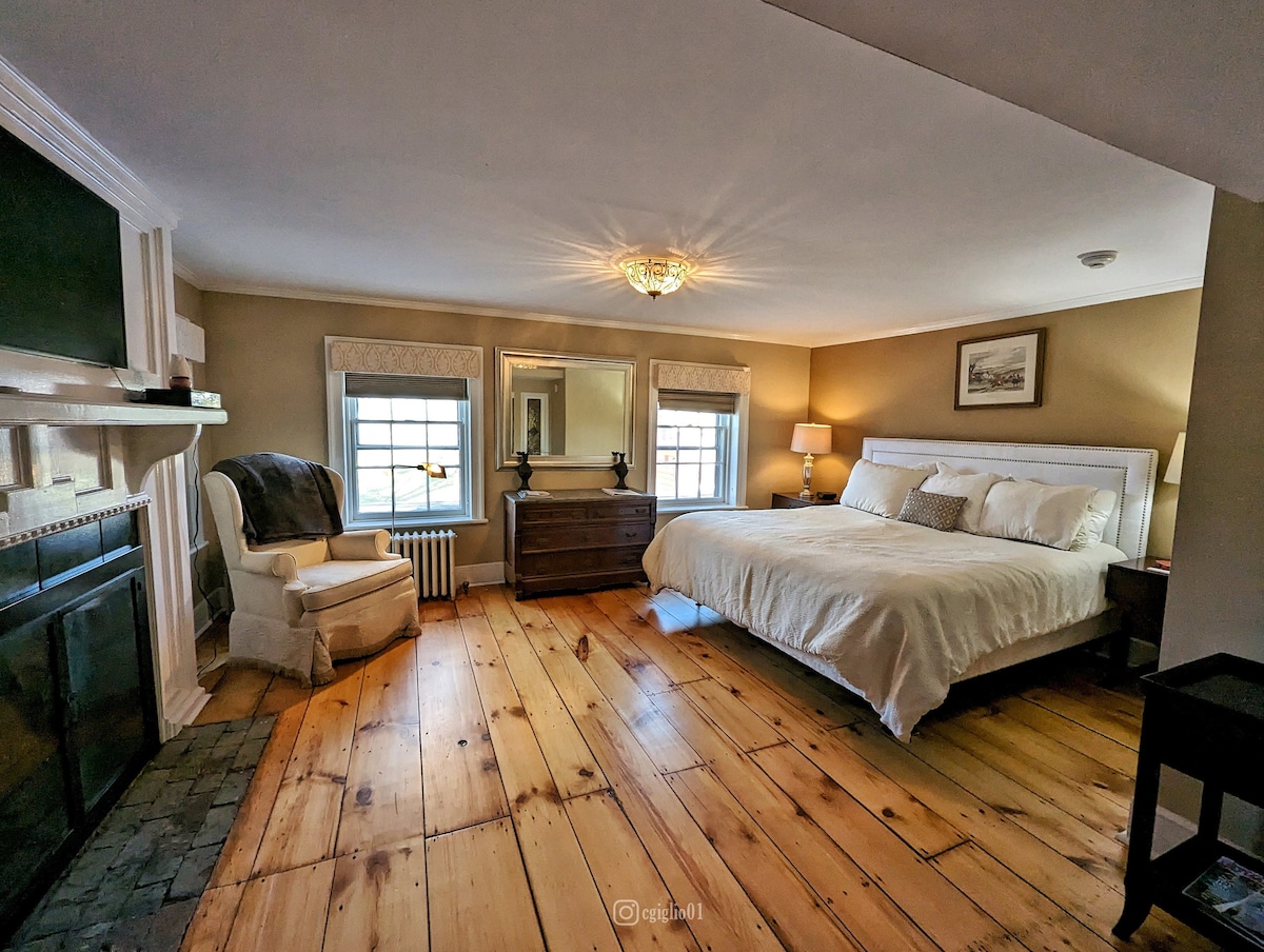 An elegantly furnished room features a spacious King bed, stylish dresser, and a lounge chair. Sunlight fills the space through multiple windows. A marble fireplace and wooden floor enhance the warm, inviting ambiance.