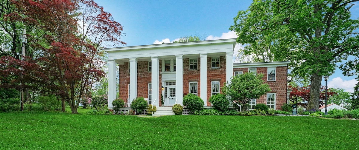 The grand exterior of the house features a classic design with large white columns and a red brick facade. Lush greenery surrounds the property, complemented by flowering trees and well-maintained bushes, creating a serene and inviting atmosphere.