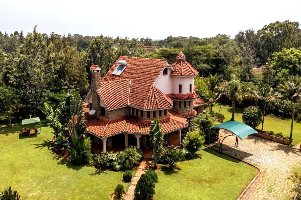 The image presents a spacious modern house with a terracotta roof situated amid lush greenery. Surrounding gardens feature palm trees and well-maintained lawns, enhancing a scenic and tranquil atmosphere. A paved driveway leads to the entrance, providing accessible access to the residence.