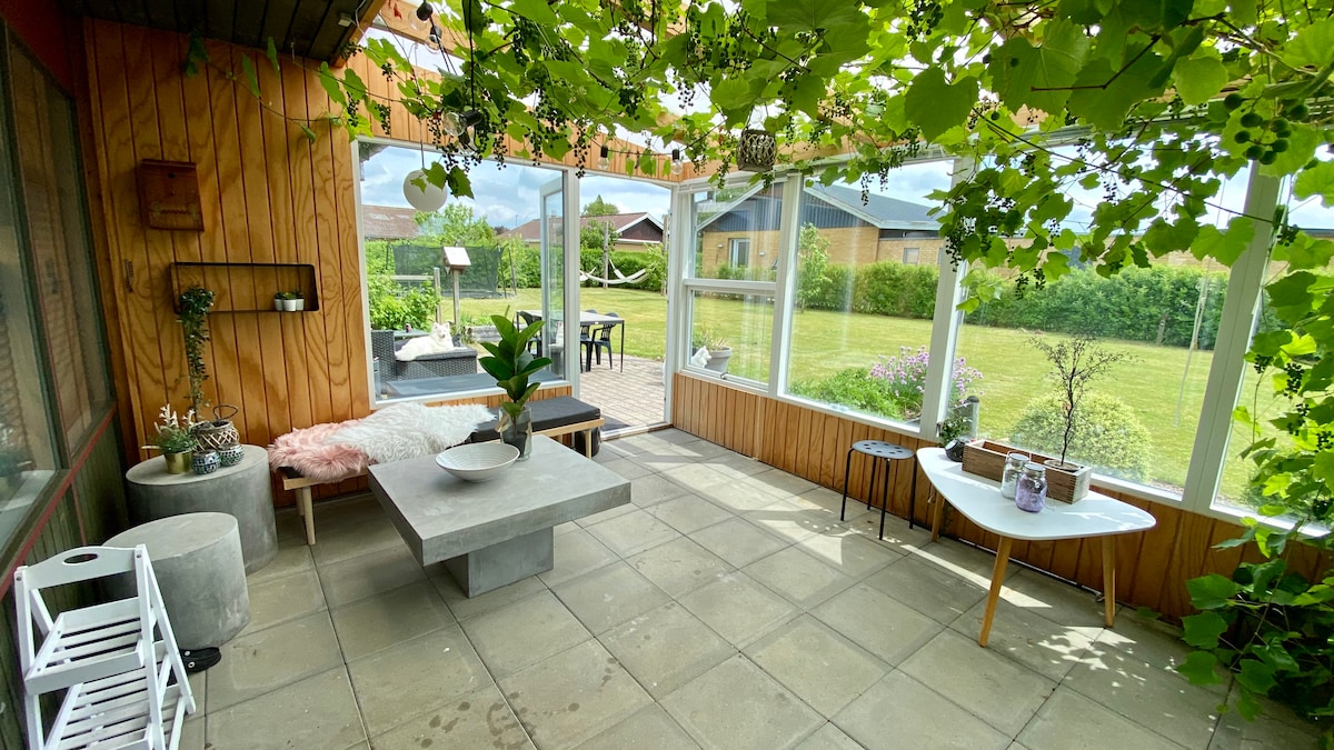 A bright sunroom is enclosed by large glass panels, allowing abundant natural light. A central concrete table is surrounded by varied seating options, including a chic white table. Lush greenery frames the space, and a well-maintained garden is visible through the glass.