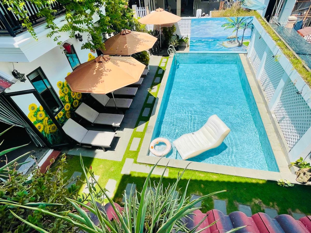 A serene outdoor area features a clear swimming pool surrounded by lounge chairs and umbrellas. Lush greenery and a mural depicting palm trees enhance the space. A shaded seating area is positioned near the pool, inviting relaxation in a calm environment.