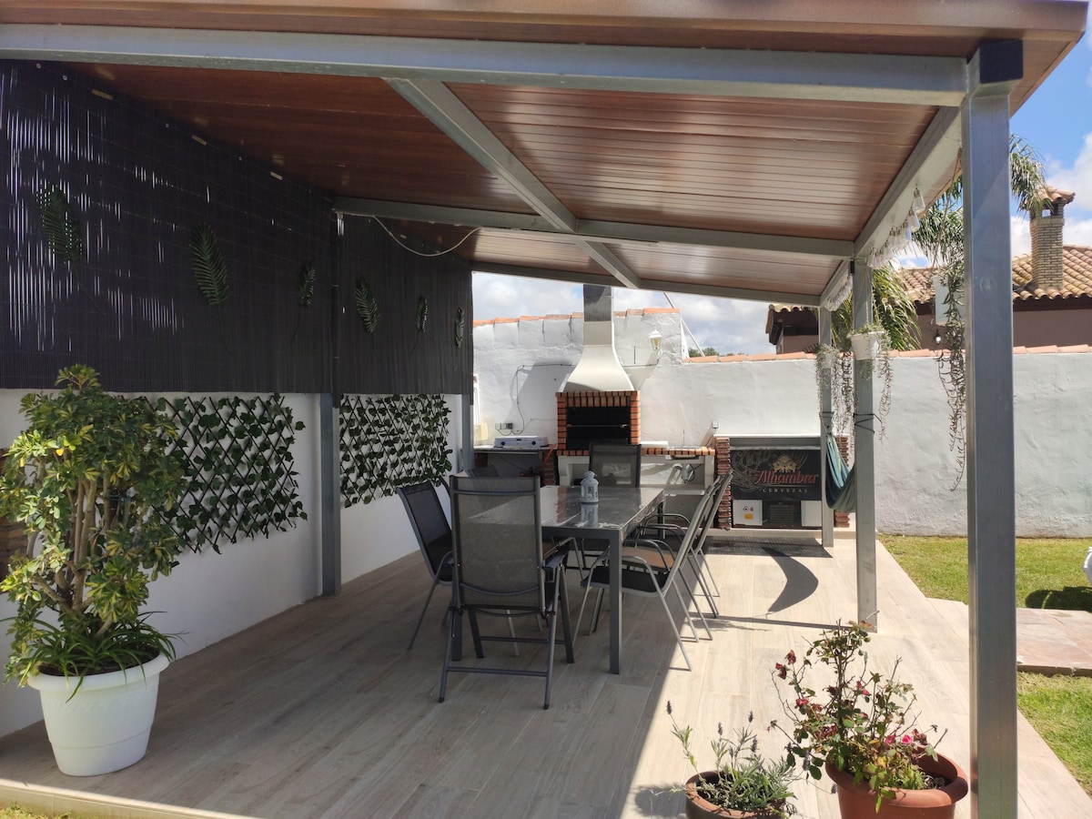 An outdoor dining area is presented under a covered terrace, featuring a large table surrounded by several metal chairs. The space is enhanced by a wall of greenery and includes a grill positioned against the back wall. Potted plants add a touch of nature.