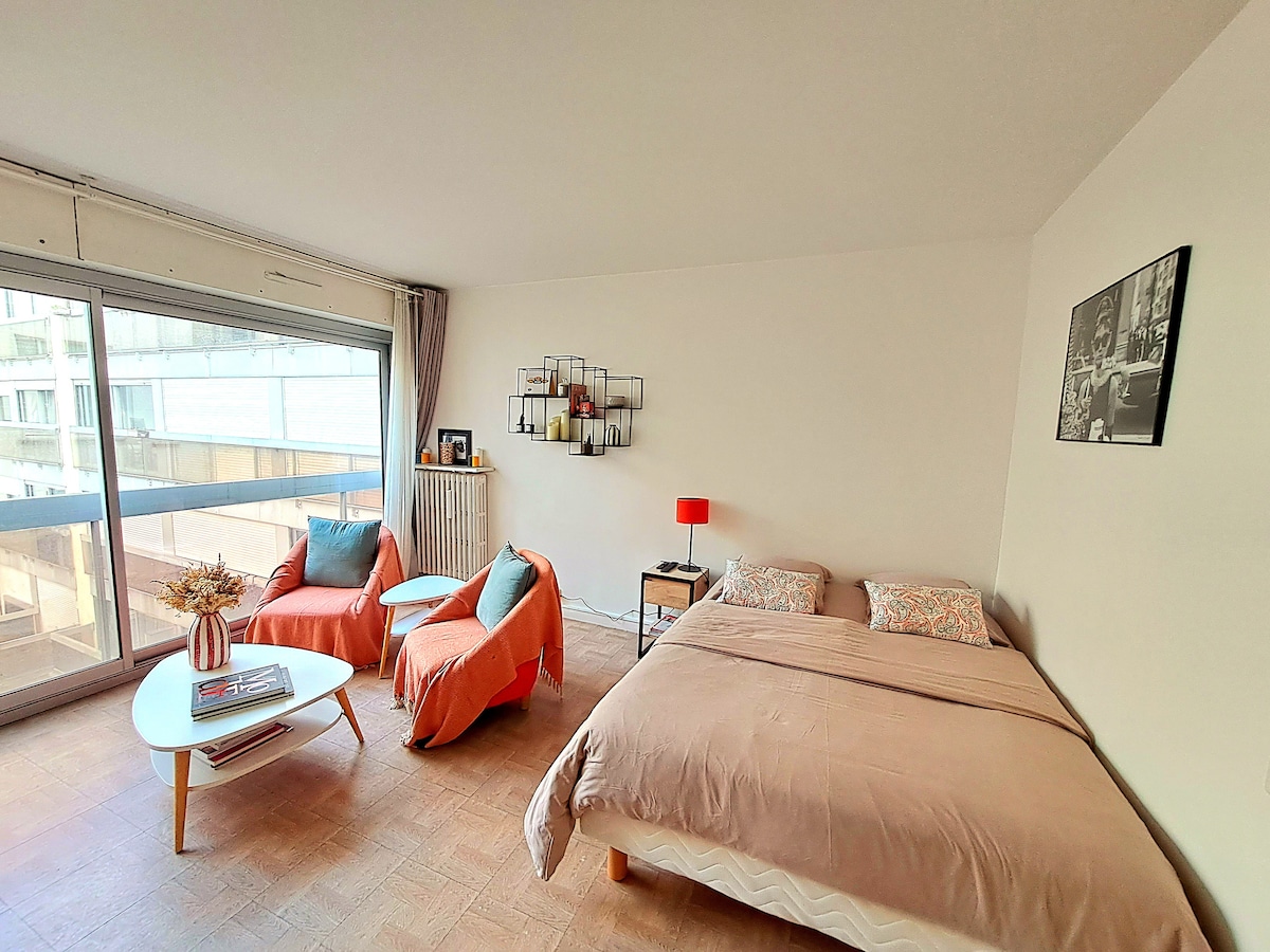 A well-lit main area features a spacious bed with soft linens and two accent chairs in a muted hue. A low coffee table sits in front, complemented by a decorative wall piece. Large windows provide natural light and access to a balcony.