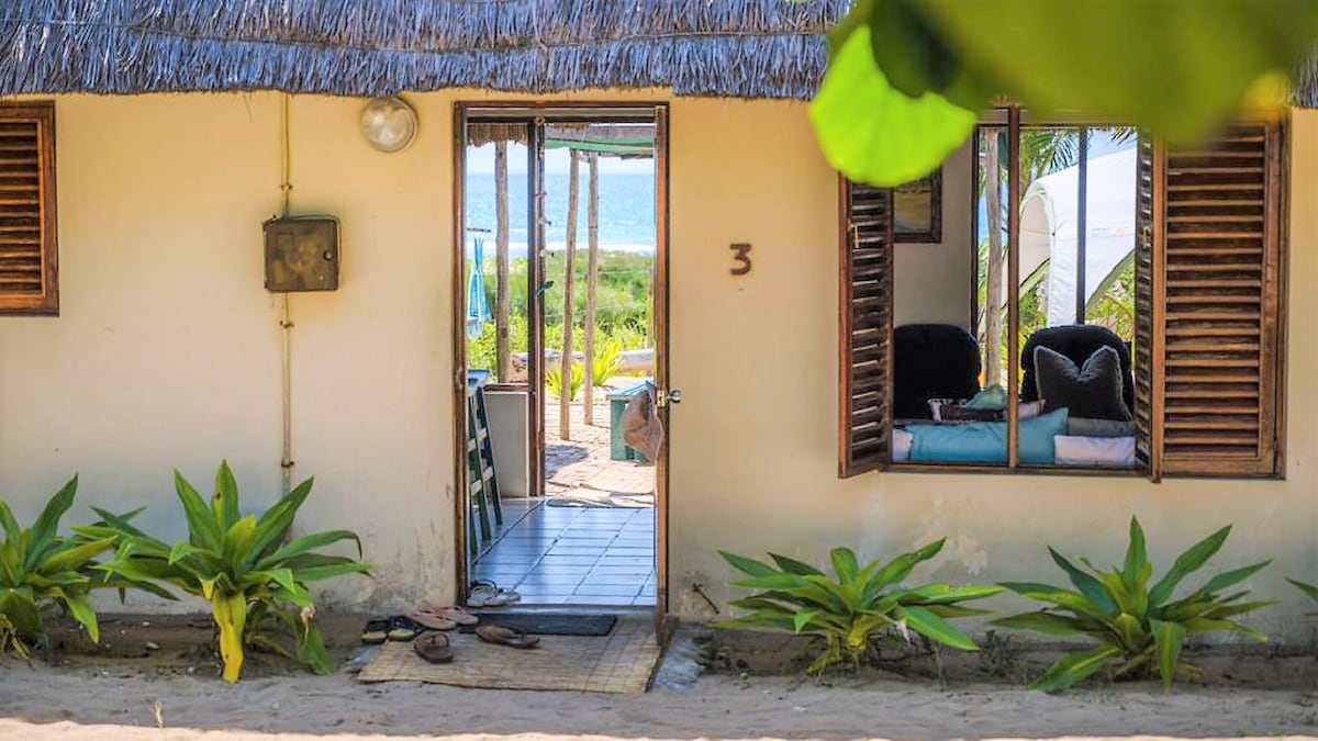 A rustic cottage entry is showcased, featuring a wooden door and shuttered windows. Lush green plants line the sandy pathway leading in. Through the doorway, glimpses of the beach and azure waters can be seen, inviting a relaxed coastal atmosphere.