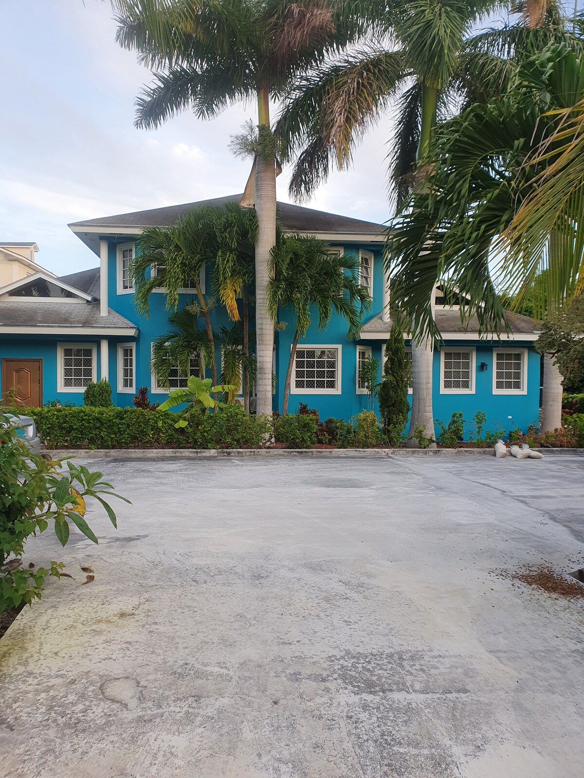 A vibrant blue house is surrounded by lush green palm trees and shrubbery. The two-story structure features several windows and a welcoming entrance. The driveway is spacious and leads to the front door, while the landscape is well-maintained, offering a tropical feel.