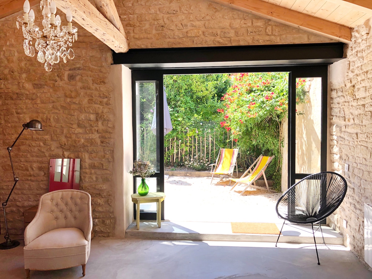 An inviting space is characterized by exposed stone walls and a wooden ceiling. A large glass door opens to a garden area with vibrant greenery. A comfortable chair and a small table are positioned nearby, alongside a stylish lamp and a decorative chandelier providing soft lighting.