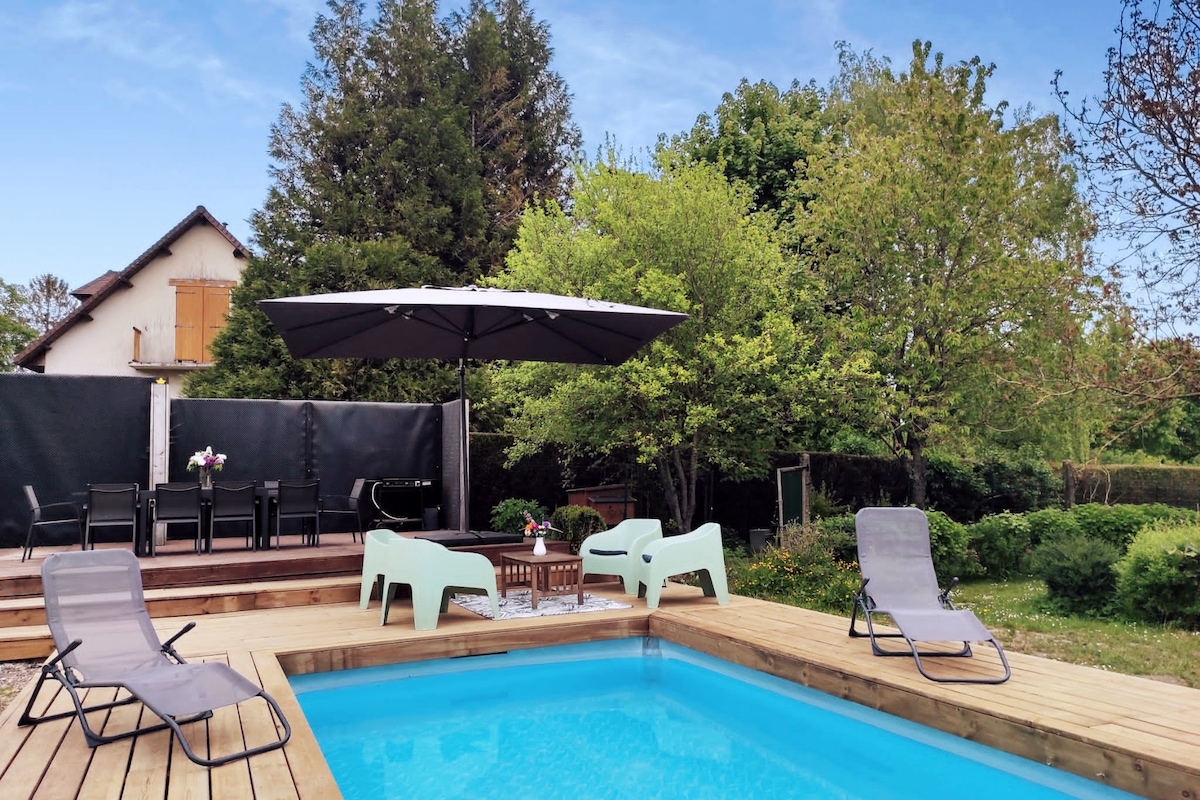 A wooden deck surrounds a sparkling pool, complemented by several lounge chairs and a large umbrella for shade. Lush greenery is visible in the background, enhancing the outdoor setting. A dining area is also present, positioned behind the pool area.