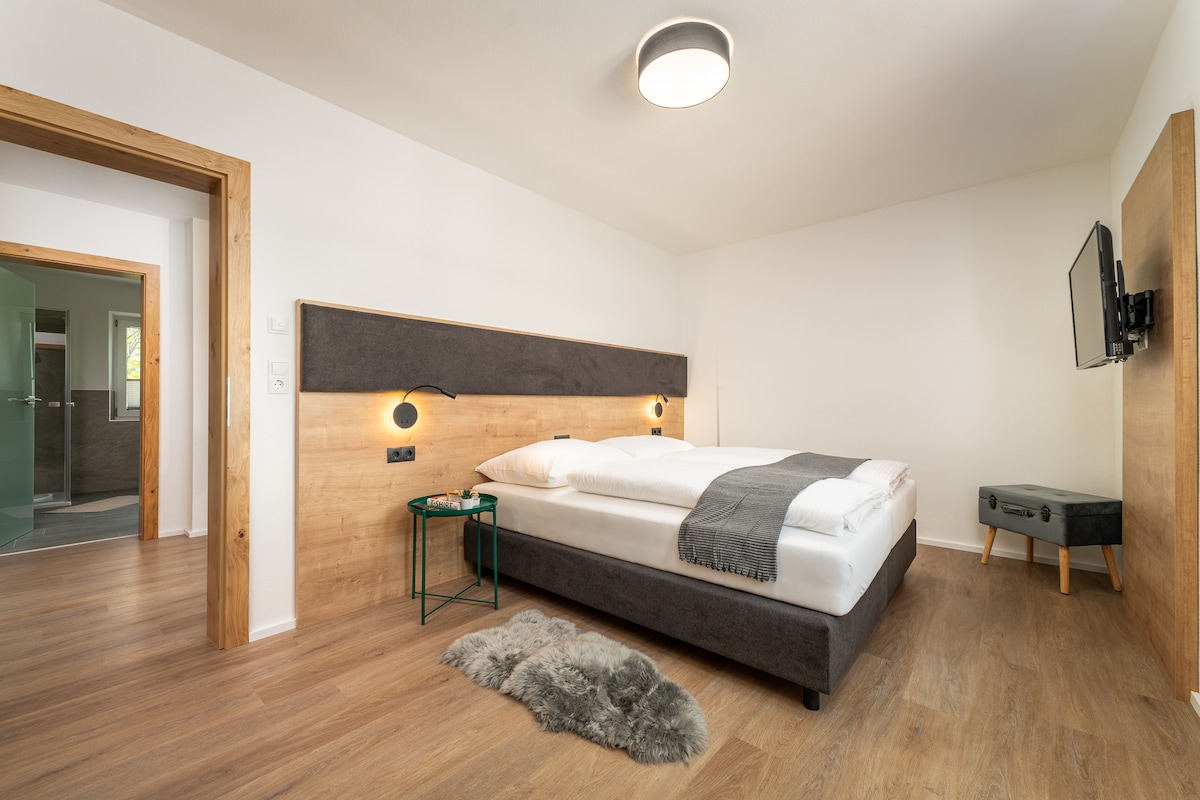 A spacious bedroom is equipped with a large bed featuring a neutral-colored headboard. A small green table sits beside the bed, and a cozy throw lies beneath it. Warm wood flooring complements the modern design, and a wall-mounted television is visible next to the door leading to the bathroom.