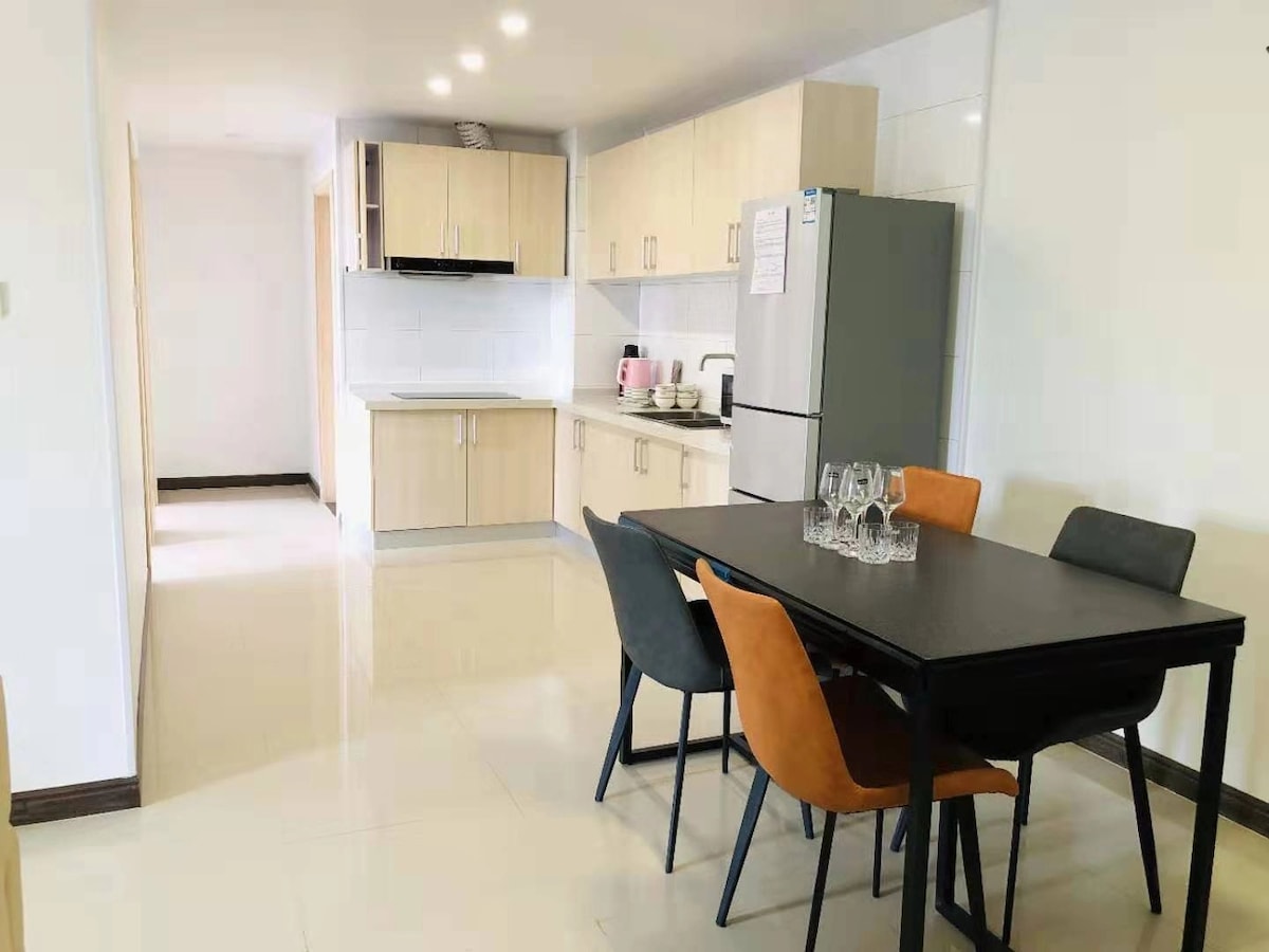 A bright and functional kitchen area features modern cabinets and a sleek refrigerator. A dining table with four chairs is positioned to one side, complemented by a set of glasses placed in the center. Natural light enhances the space, highlighting the clean tile flooring.