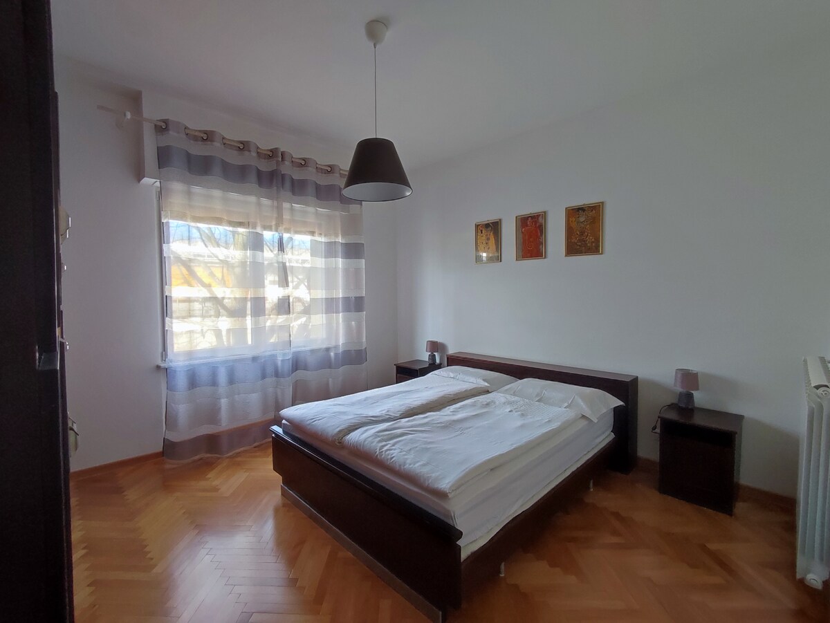 A bright bedroom features a spacious bed with crisp white bedding. Light streams in through a large window adorned with sheer curtains. Two bedside tables with lamps are positioned beside the bed. Three framed art pieces hang on the wall, adding subtle color.