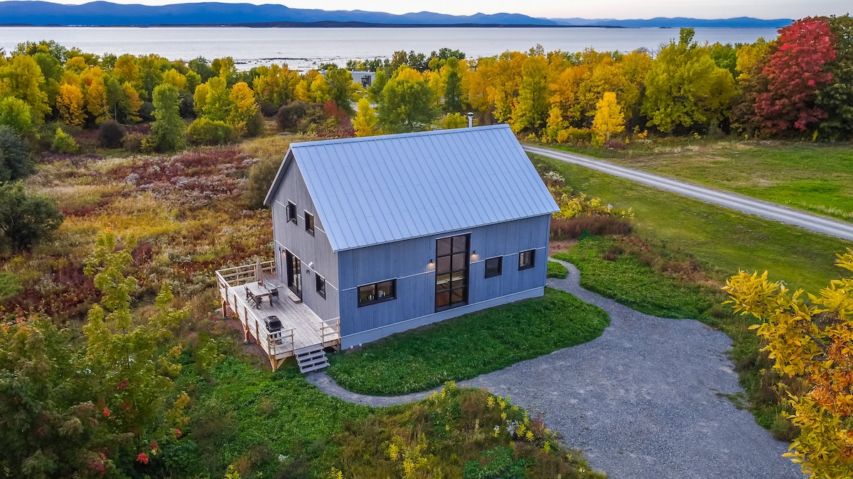 The exterior of the house displays a modern design with a silver metal roof and large central windows. Surrounded by colorful autumn foliage, the property is set against a backdrop of rolling hills and a serene body of water, suggesting an inviting atmosphere for guests.