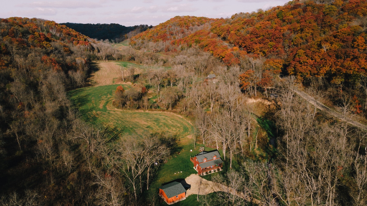 An aerial view reveals a secluded cabin nestled in a vibrant valley, surrounded by rolling hills adorned with autumn foliage. The spacious lawn is framed by trees, showcasing the natural beauty of the property and its tranquil setting.