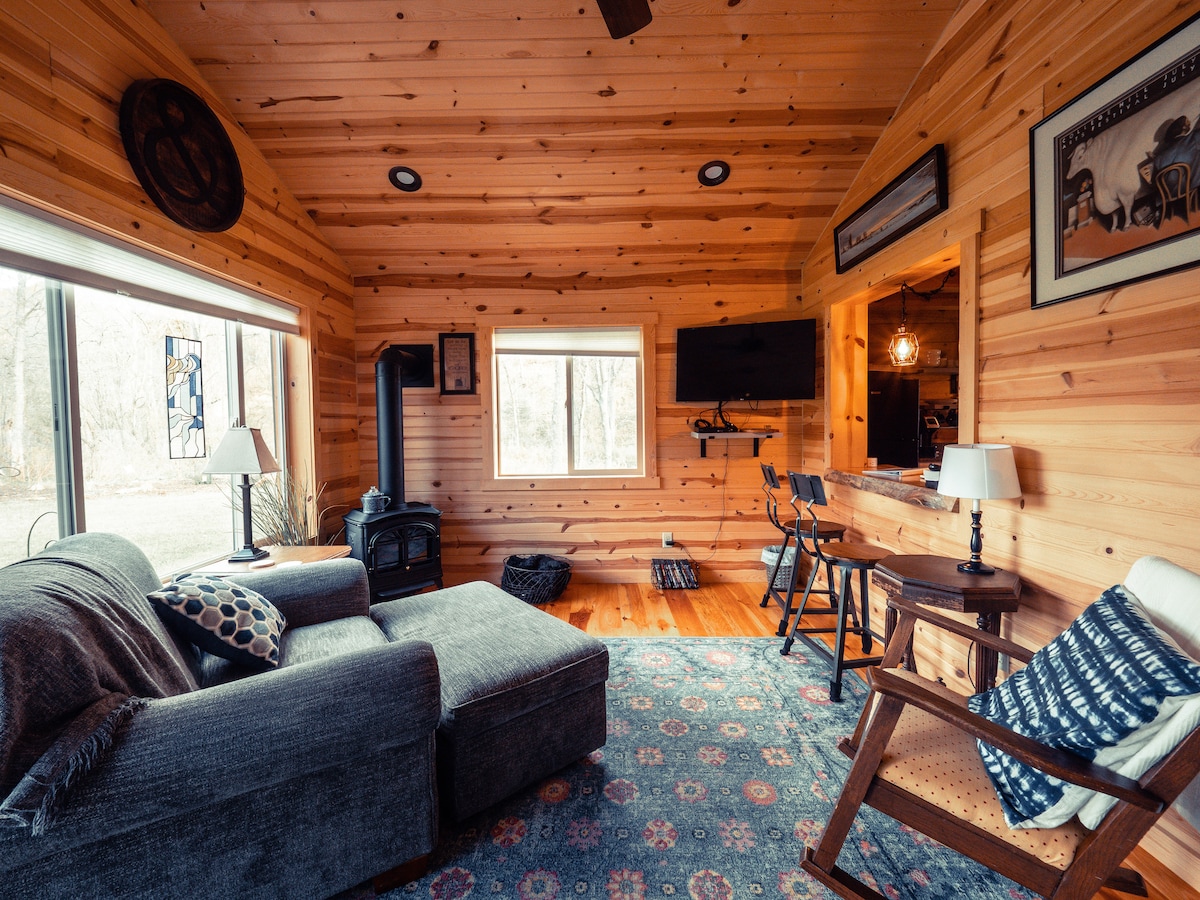 The living area features a cozy couch and a patterned rug, surrounded by wooden walls that add warmth. A large window allows natural light to illuminate the space. A TV is mounted on the wall opposite the seating. Bar stools are positioned near the kitchen area.