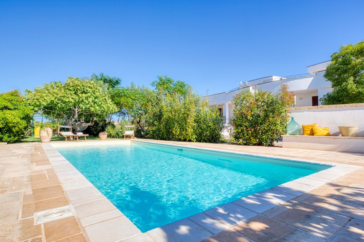 A crystal-clear swimming pool is set amidst lush greenery, featuring a spacious deck and surrounding plants. Sun loungers are positioned nearby, providing a tranquil space for relaxation. The blue sky above complements the serene outdoor setting.