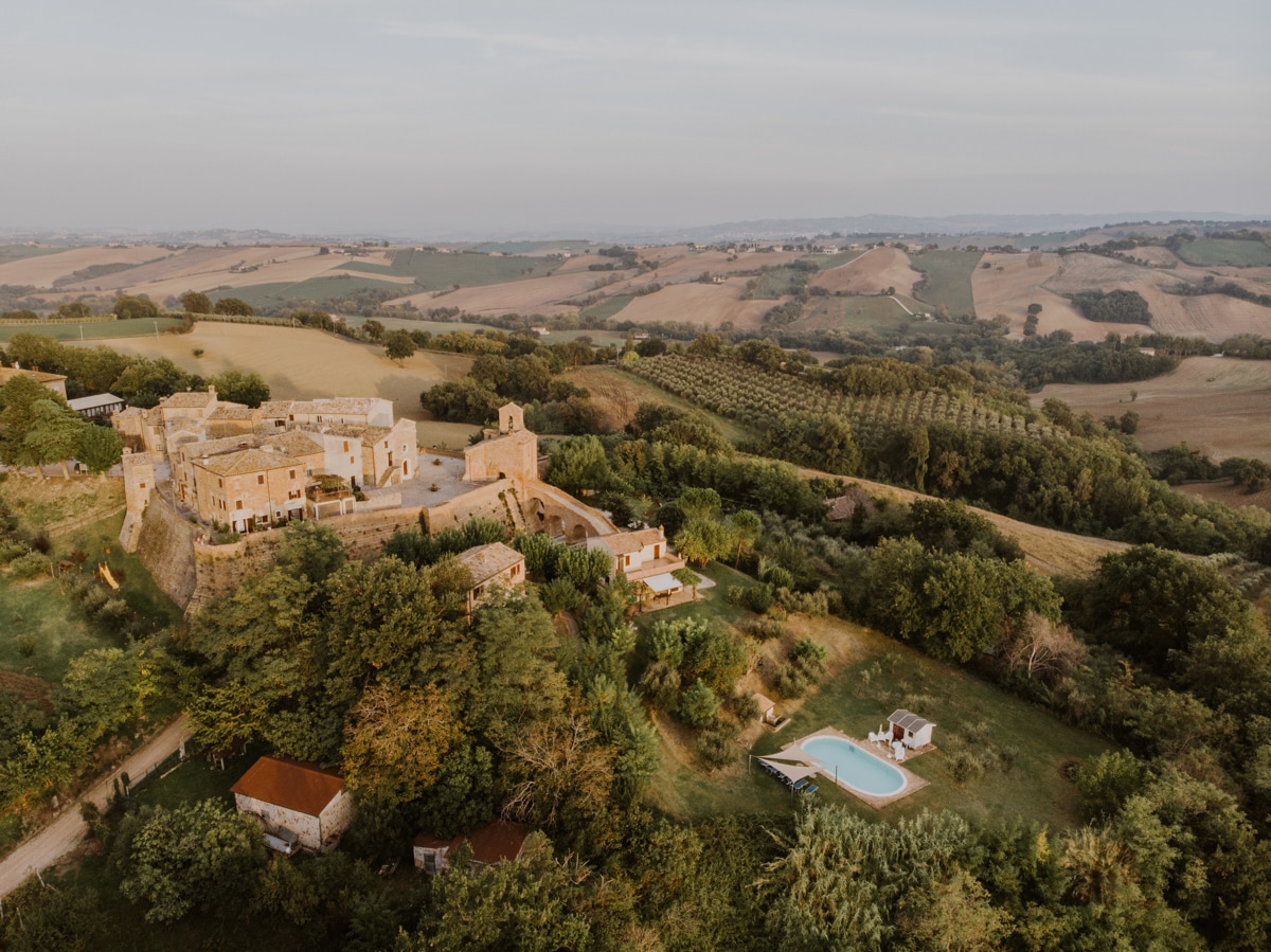 A sweeping aerial view captures a historic stone structure nestled among lush greenery, accompanied by a serene outdoor pool. Rolling hills stretch into the distance, showcasing the picturesque landscape of the surrounding countryside.