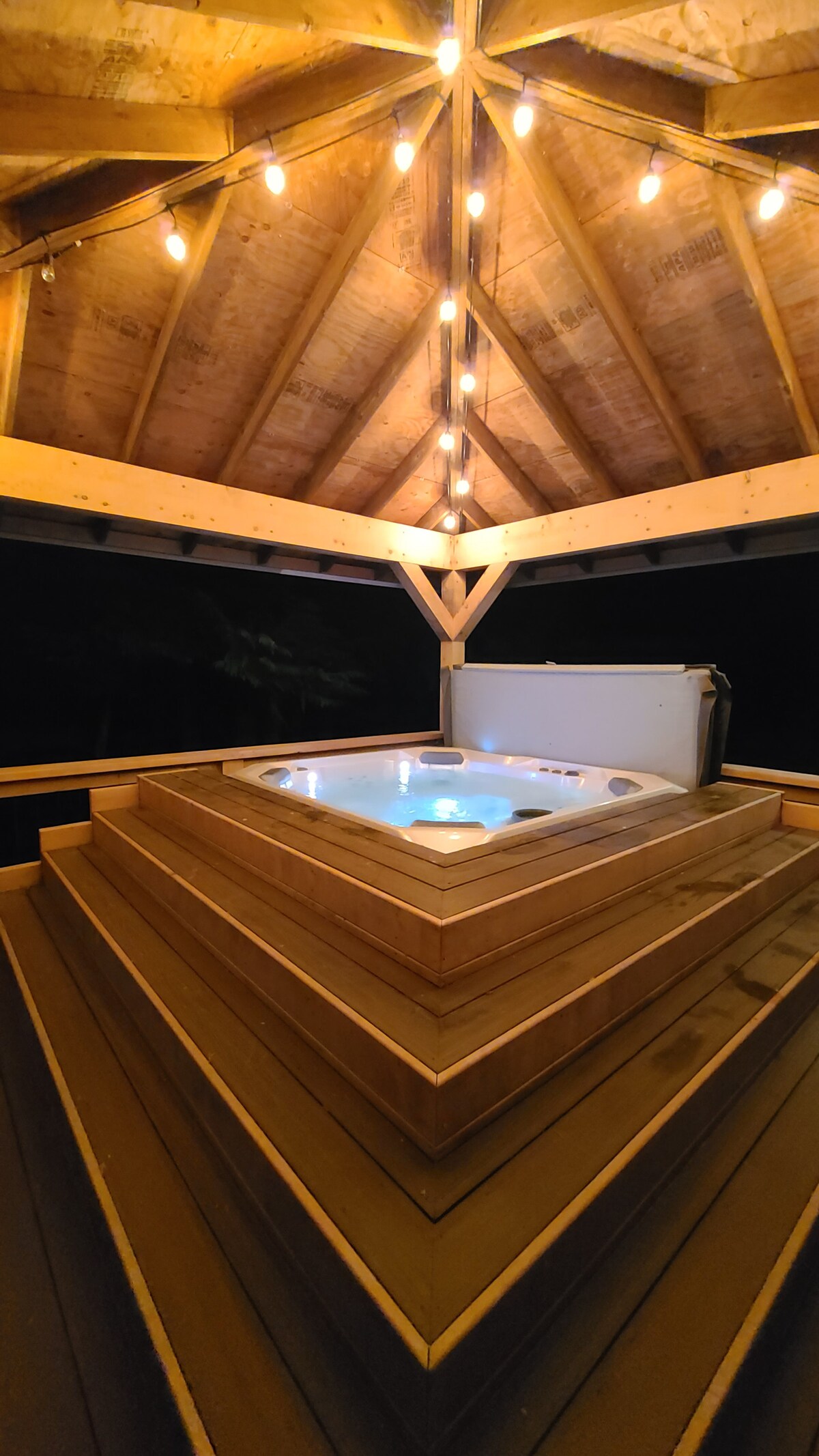 A covered hot tub rests on a multi-tiered wooden deck, illuminated by string lights above. The warm glow creates a relaxing ambiance, highlighting the clean, inviting space designed for up to six people.