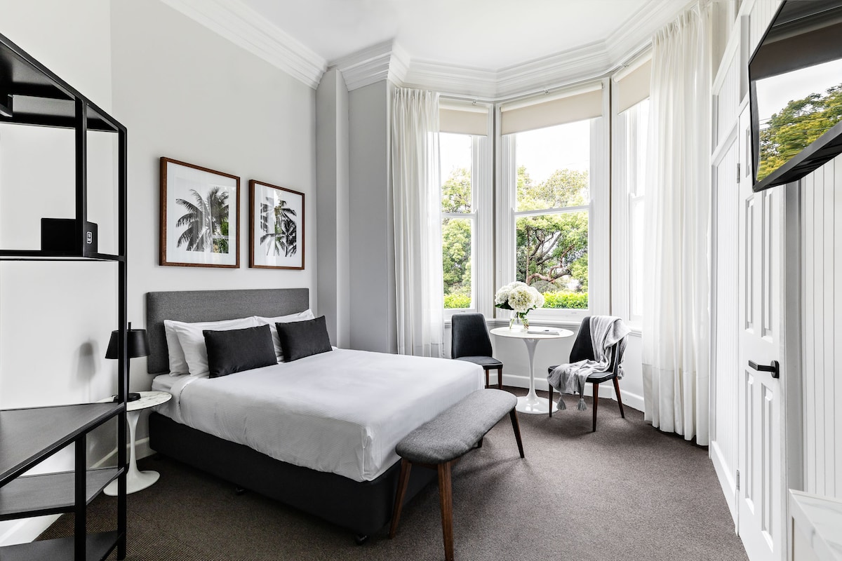A spacious guest room features a queen bed positioned under bright windows with views of greenery. Two framed botanical prints adorn the wall, complementing a small dining table with chairs, and a stylish black shelving unit is present.