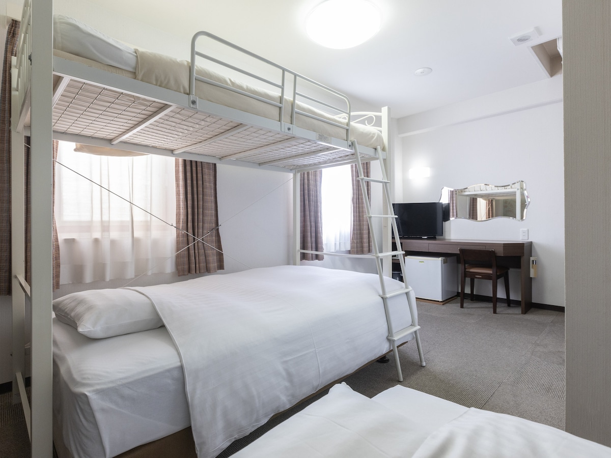 This triple room features two single beds and a bunk bed, providing ample sleeping space. Natural light filters through the sheer curtains, illuminating a flat-screen TV and a desk area. A mirror and a refrigerator are also present, enhancing the room's functionality.
