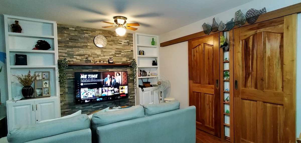 A living area features a comfortable sectional sofa facing a wall-mounted 55" smart TV. Accent shelves hold decorative items, and warm wooden sliding doors complement the stone wall. A ceiling fan is visible, adding to the inviting atmosphere of the space.