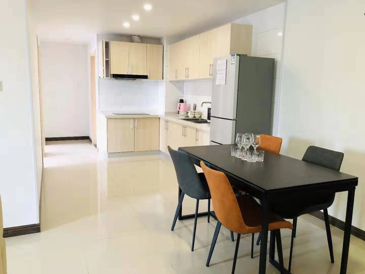 A modern kitchen and dining area feature light wooden cabinetry and a sleek countertop. A dining table with four chairs is positioned near the kitchen, enhancing the open layout. A refrigerator and various kitchen appliances are visible, contributing to a functional space.