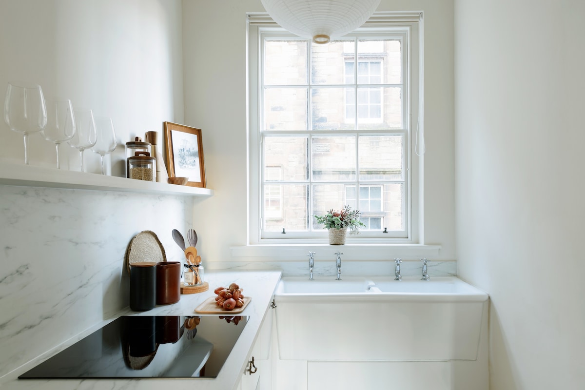 A well-appointed kitchen features a marble countertop, an original butler sink, and modern cooking appliances. Glassware is neatly arranged on the shelf, while a decorative frame and a small potted plant add a touch of warmth. Natural light floods in through the large window.